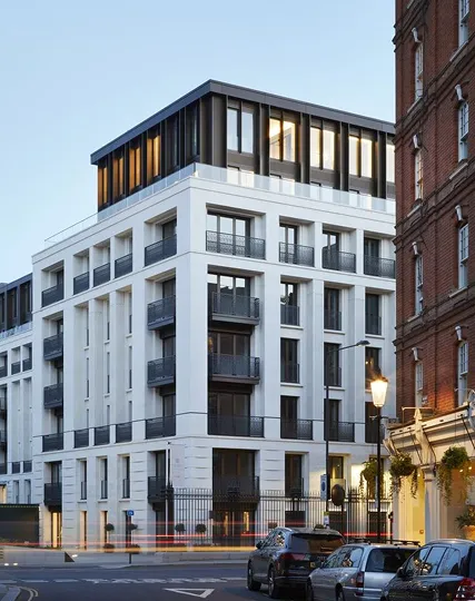 Modern multi-story white building with black window frames and balconies at dusk, street lamps and parked cars in front.