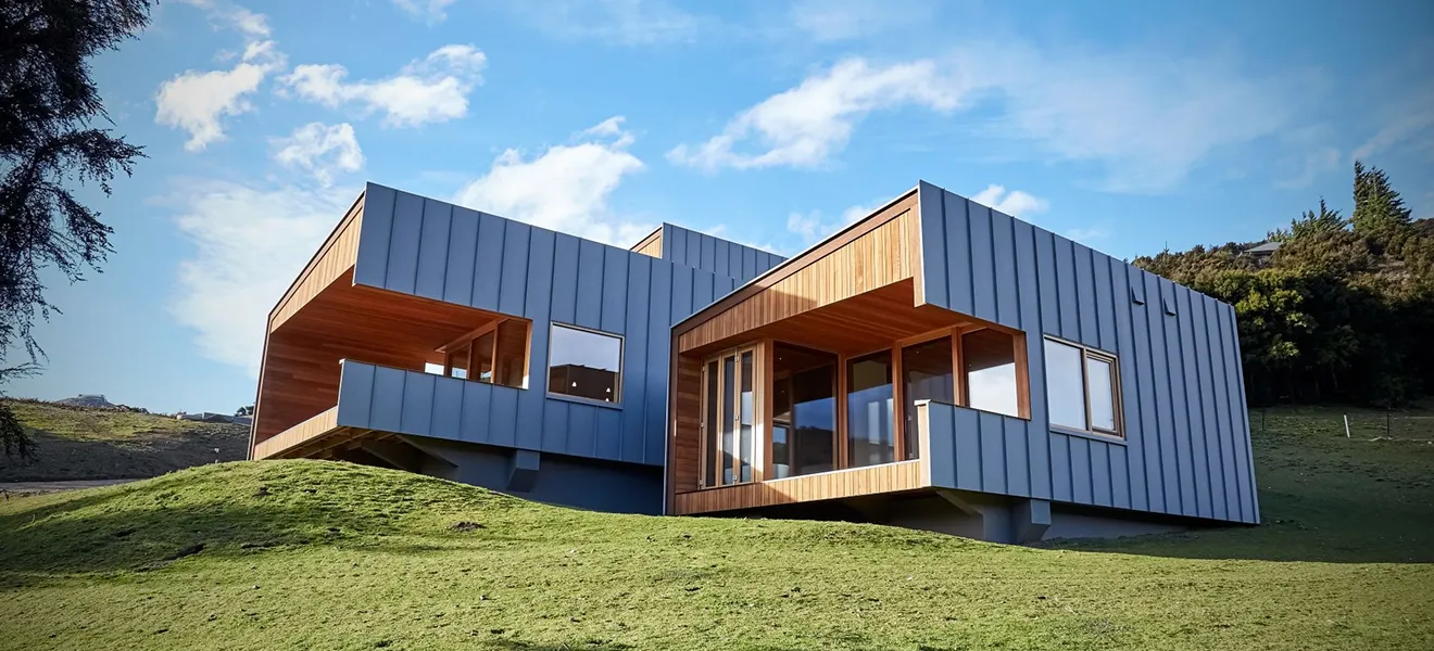 Modern house with gray metal siding and wooden accents built on a grassy hillside under a blue sky.