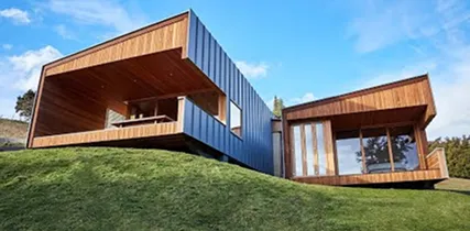Modern house with cantilevered wooden sections and large glass windows on a grassy hill under blue sky.